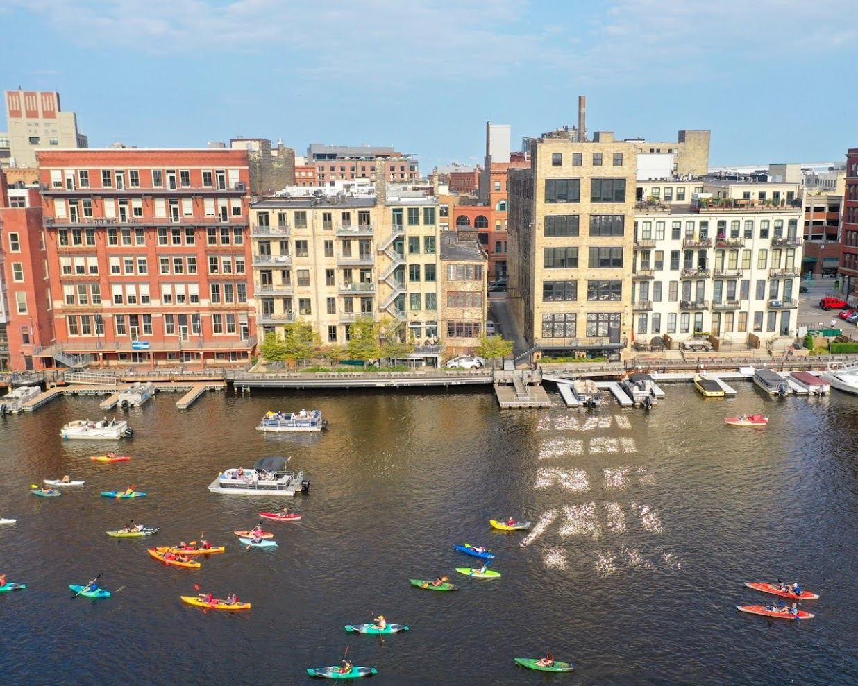 Aerial view of the Milwaukee River curving past the Third Ward with boats moored along the Riverwalk and the downtown skyline behind.