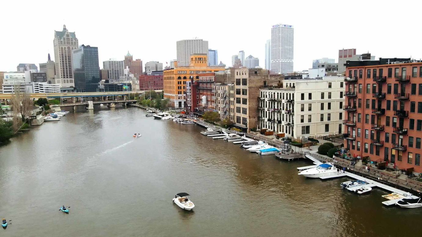 Wide view of the Third Ward riverfront with the Milwaukee skyline behind and boats docked along the Riverwalk.