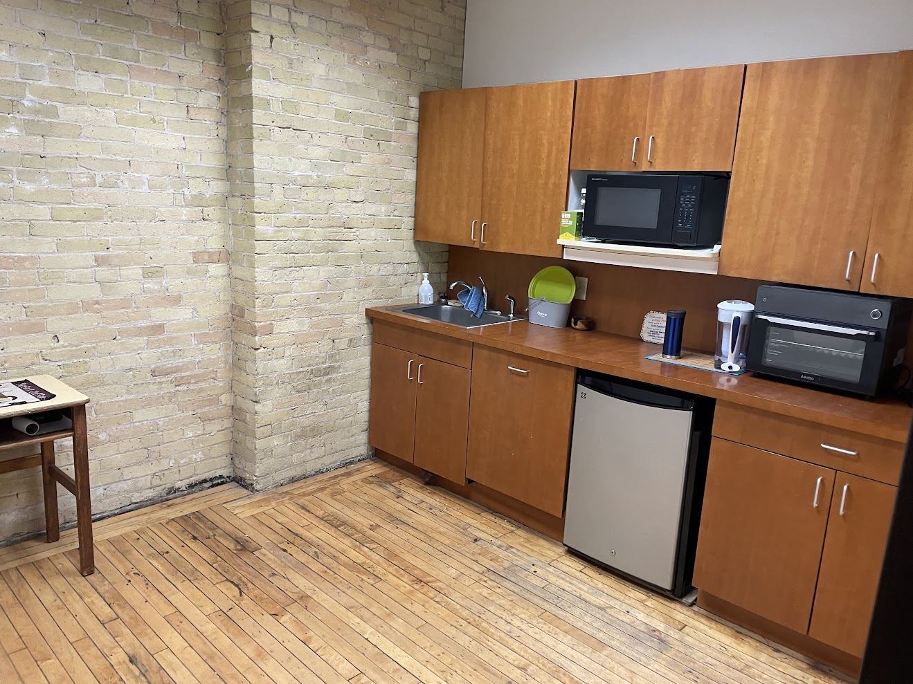 Suite 200 kitchen with wood cabinetry, sink, under-counter refrigerator and exposed brick wall.
