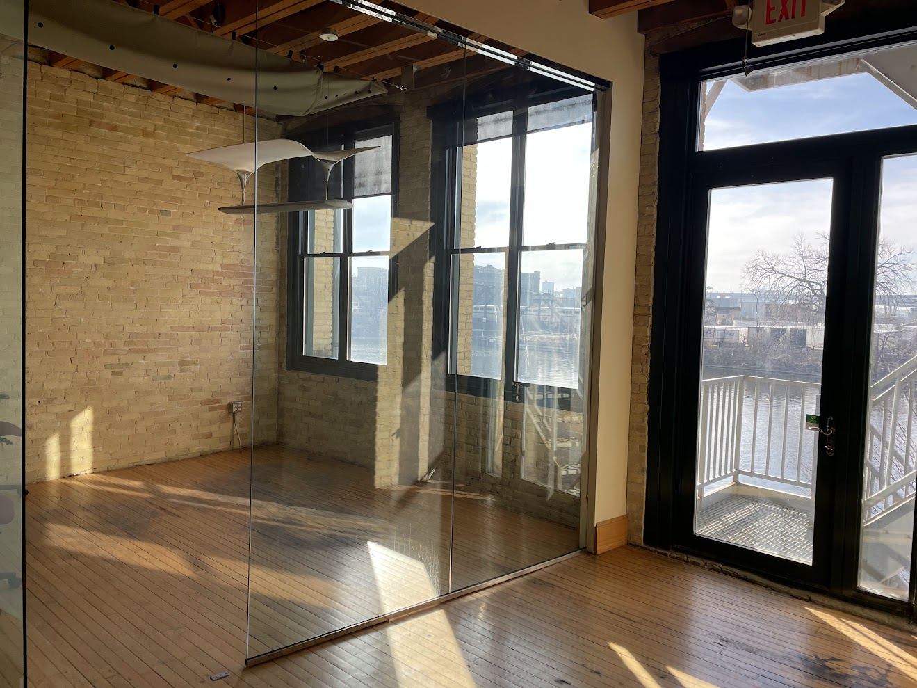 Glass-walled corner office in Suite 250 with exposed Cream City brick, timber ceiling and tall windows, beside doors opening onto a river-facing landing.