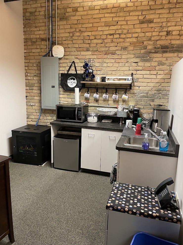 Kitchenette in Suite 400 with sink, microwave, under-counter refrigerator and a mug rack against exposed brick.